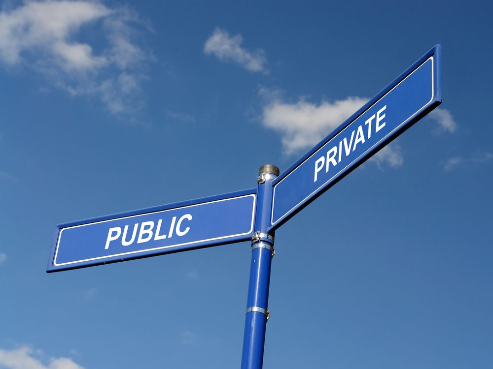 A blue street sign shows the word Private pointing in one direction and the word Public pointing in another direction. The background shows clouds, and the street sign is pictured from below.